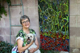 A woman holding a small dog, kneeling in a garden next to a colorful mosaic.